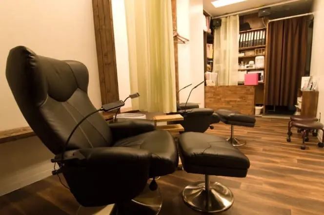 落ち着いた照明と木目調の床が印象的な、リクライニングチェアを備えた施術スペース。まつげエクステやネイルなどの美容サロンを思わせる個室風のインテリア。