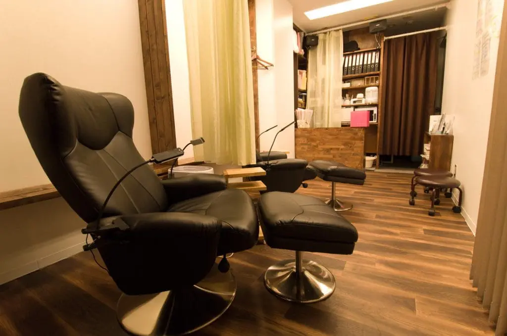 落ち着いた照明と木目調の床が印象的な、リクライニングチェアを備えた施術スペース。まつげエクステやネイルなどの美容サロンを思わせる個室風のインテリア。