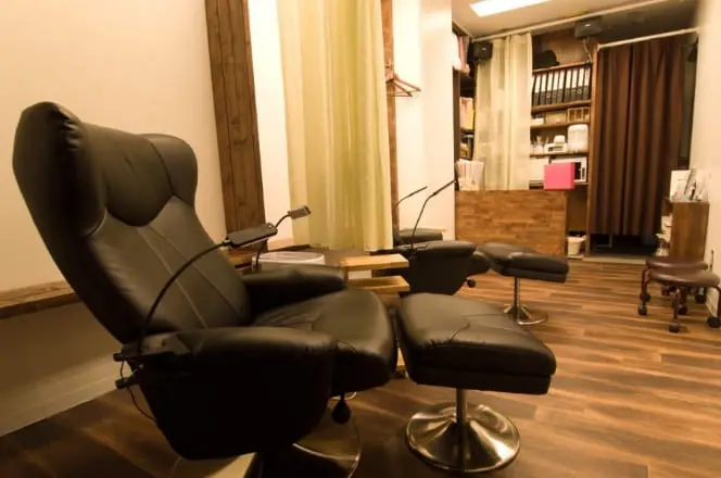 落ち着いた照明と木目調の床が印象的な、リクライニングチェアを備えた施術スペース。まつげエクステやネイルなどの美容サロンを思わせる個室風のインテリア。